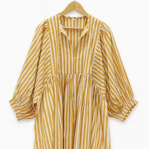 May include: A long, yellow and white striped dress with long sleeves and a collared neckline. The dress has a loose, flowing fit and is hanging on a wooden hanger against a white background.