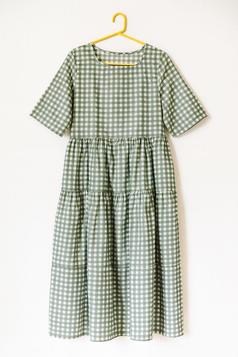 Puede incluir: Vestido midi a cuadros verdes y blancos con mangas cortas y falda escalonada. El vestido est&aacute; colgado de una percha amarilla sobre un fondo blanco. El vestido es de tejido ligero.