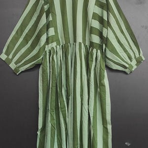 May include: A green and light green striped dress with a round neckline and three-quarter sleeves. The dress has a gathered waist and a loose, flowing fit. The fabric appears to be lightweight and suitable for warmer weather. The dress is hanging on a wooden hanger.