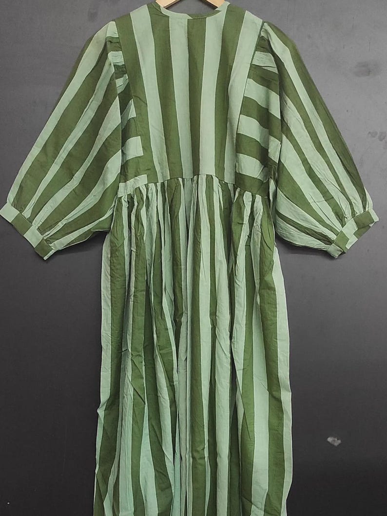 May include: A green and light green striped dress with a round neckline and three-quarter sleeves. The dress has a gathered waist and a loose, flowing fit. The fabric appears to be lightweight and suitable for warmer weather. The dress is hanging on a wooden hanger.