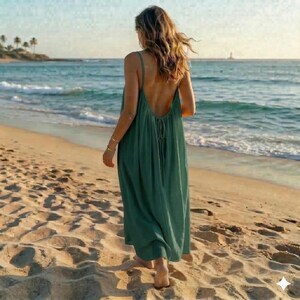 May include: A long, flowing, backless, emerald green dress. The dress has thin straps and a low back, and is being worn on a sandy beach. The person wearing the dress is walking towards the ocean, with the waves in the background.