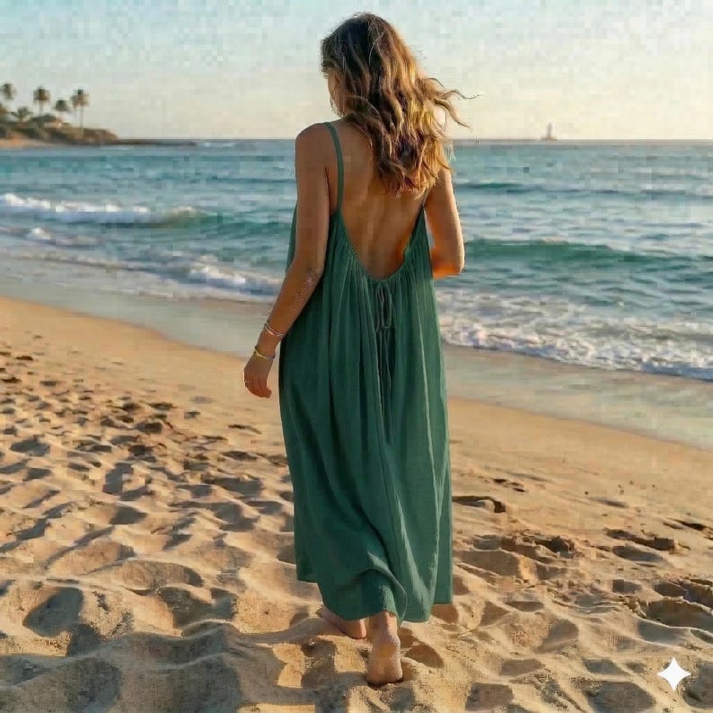 May include: A long, flowing, backless, emerald green dress. The dress has thin straps and a low back, and is being worn on a sandy beach. The person wearing the dress is walking towards the ocean, with the waves in the background.
