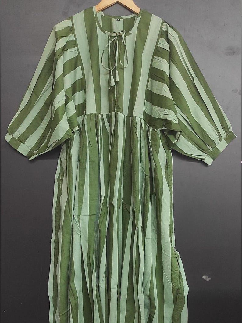 May include: A green and light green striped dress with a round neckline and three-quarter sleeves. The dress features a tie detail at the neckline and a gathered skirt. The fabric appears to be lightweight, suitable for warmer weather.