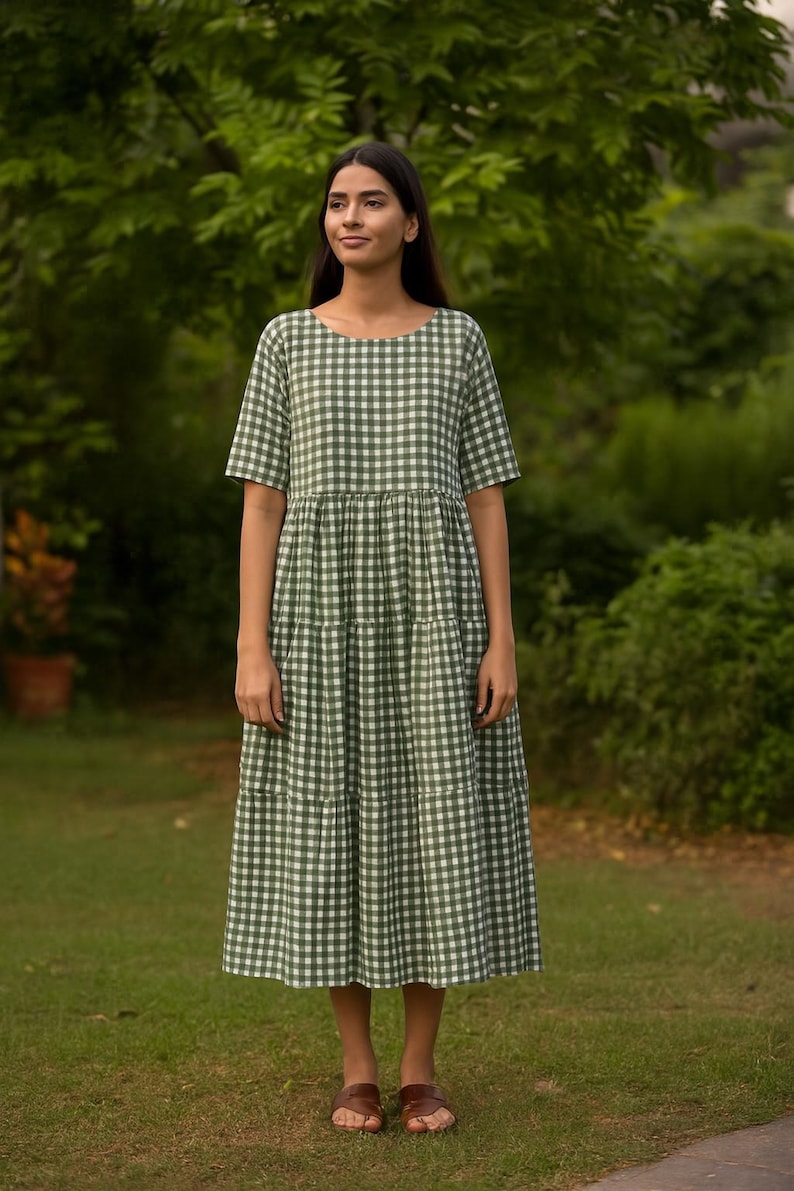 Puede incluir: Vestido midi con estampado de cuadros vichy verde y blanco, con mangas cortas. El vestido tiene cuello redondo y falda escalonada. La persona lleva sandalias marrones. El fondo es un c&eacute;sped verde y &aacute;rboles.