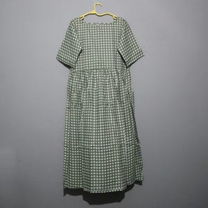 Puede incluir: Un vestido de patr&oacute;n a cuadros verdes y blancos con mangas cortas y cintura fruncida. El vestido est&aacute; colgado de una percha amarilla sobre un fondo gris. El vestido es de largo midi.
