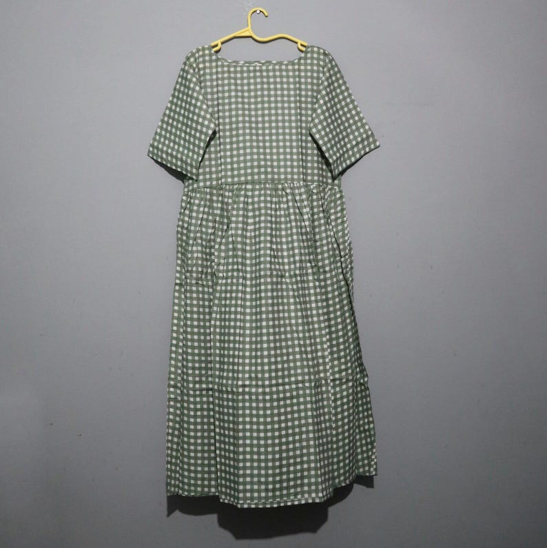 Puede incluir: Un vestido de patr&oacute;n a cuadros verdes y blancos con mangas cortas y cintura fruncida. El vestido est&aacute; colgado de una percha amarilla sobre un fondo gris. El vestido es de largo midi.