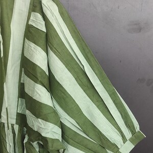 May include: Close-up of a green and white striped garment. The fabric appears to be lightweight, with vertical stripes running the length of the sleeve and body. The sleeve has a gathered, puffy design at the shoulder and a buttoned cuff.
