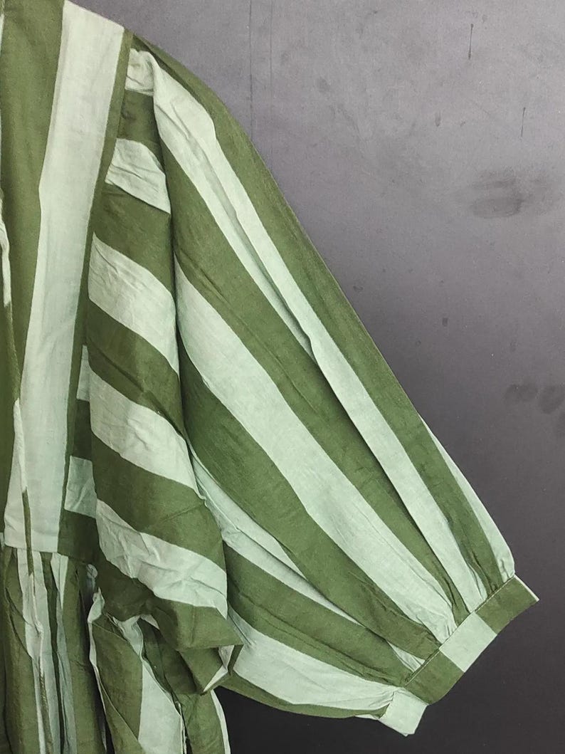 May include: Close-up of a green and white striped garment. The fabric appears to be lightweight, with vertical stripes running the length of the sleeve and body. The sleeve has a gathered, puffy design at the shoulder and a buttoned cuff.