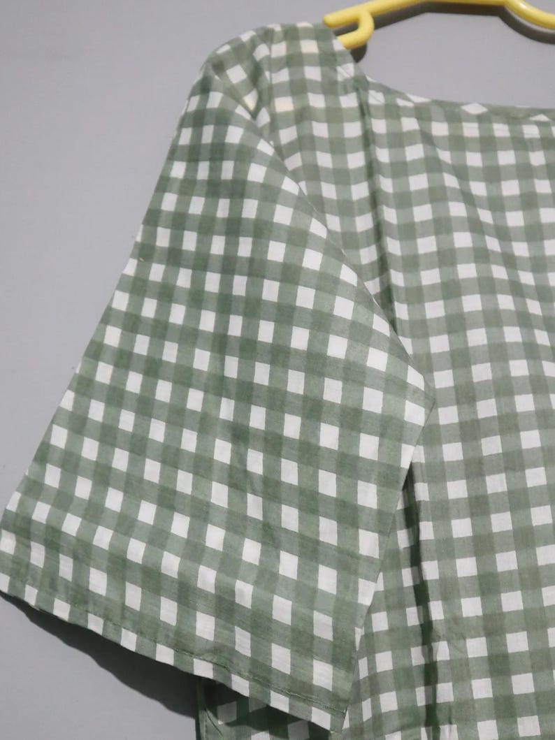 Puede incluir: Top a cuadros verdes y blancos con mangas cortas. La tela es un material ligero y transpirable, y el patr&oacute;n es un dise&ntilde;o de cuadros cl&aacute;sico. La parte superior est&aacute; colgada de una percha amarilla.