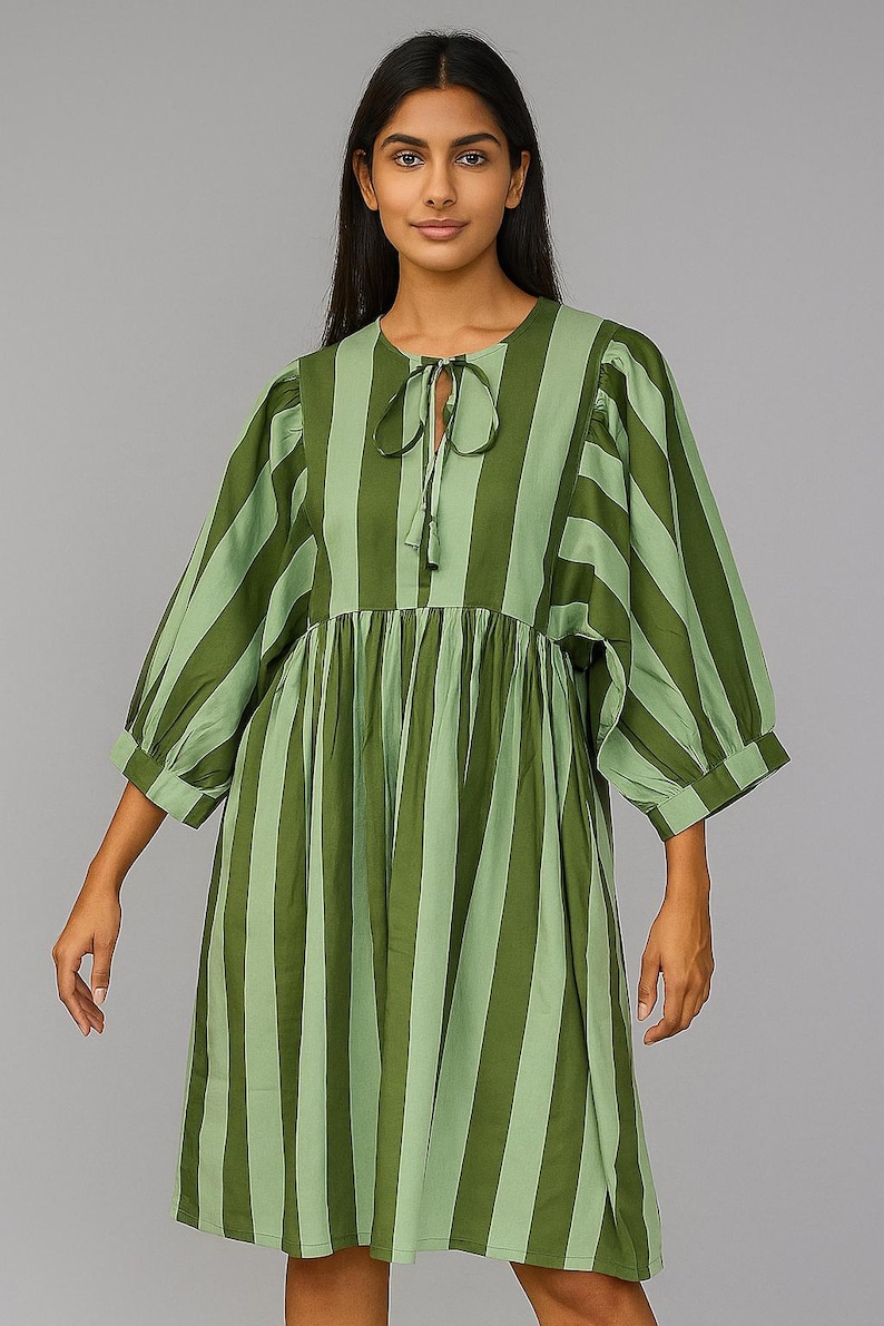May include: A green and light green striped dress with three-quarter sleeves. The dress features a tie at the neckline and a gathered waist. The model is wearing the dress against a gray background.