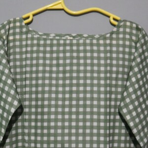 Puede incluir: Un vestido a cuadros de color verde y blanco cuelga de una percha amarilla. El vestido tiene un escote redondo y mangas cortas. La tela parece ligera, ideal para el clima c&aacute;lido. El vestido mide aproximadamente 100 cm de largo.