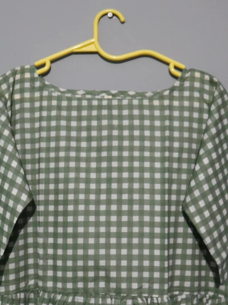 Puede incluir: Un vestido a cuadros de color verde y blanco cuelga de una percha amarilla. El vestido tiene un escote redondo y mangas cortas. La tela parece ligera, ideal para el clima c&aacute;lido. El vestido mide aproximadamente 100 cm de largo.