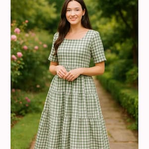Puede incluir: Un vestido de cuadros verdes y blancos con mangas cortas y falda escalonada. El vestido tiene un escote cuadrado y llega a la rodilla. La mujer est&aacute; de pie en un camino de piedra en un jard&iacute;n.
