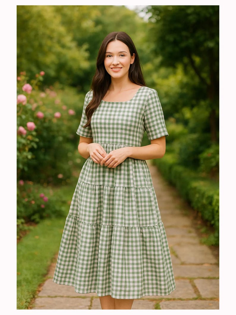 Puede incluir: Un vestido de cuadros verdes y blancos con mangas cortas y falda escalonada. El vestido tiene un escote cuadrado y llega a la rodilla. La mujer est&aacute; de pie en un camino de piedra en un jard&iacute;n.