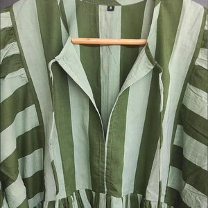 May include: A green and light green striped dress with a V-neckline and a gathered skirt. The dress is hanging on a wooden hanger. The fabric has a soft, flowing appearance.