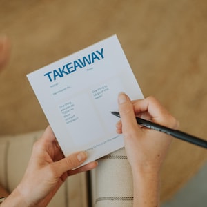 May include: A white Takeaway notepad with blue text is held by a person. The notepad has prompts for self-reflection, including "One thing you can do TODAY to show yourself kindness?" and "One thing to let go of this week?"