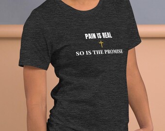 May include: Dark grey t-shirt with the text "PAIN IS REAL" above a cross symbol and the text "SO IS THE PROMISE". The shirt is made of a soft material and has short sleeves.