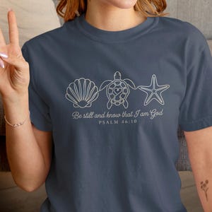 May include: A navy blue t-shirt featuring white line art of a seashell, sea turtle, and starfish. The text "Be still and know that I am God, Psalm 46:10" is printed below the graphics. The shirt is a comfortable, casual style.