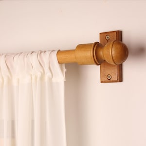 May include: A wooden curtain pole with a light finish. The pole is mounted on a wall with a wooden bracket and a decorative finial. White sheer curtains are draped on the pole.