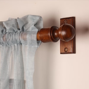 May include: A wooden curtain rod with a dark brown finish, featuring a decorative finial and a matching wall mount. Light blue sheer curtains are draped on the rod. The rod is designed for hanging curtains.