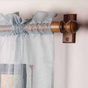 May include: A light blue sheer curtain panel with a ruffled top, hanging from a wooden rod. The rod is supported by a dark brown bracket attached to a white wall. The curtain is semi-transparent.