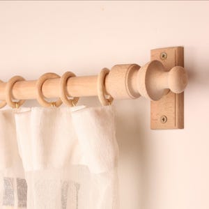 May include: A wooden curtain rod with rings and a decorative finial. The rod is mounted on a wall with a light-colored finish. The curtain rod is made of light-colored wood and holds sheer white curtains.