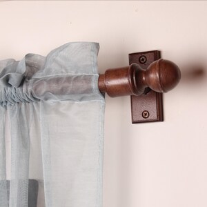 May include: A wooden curtain rod with a decorative finial and a matching bracket. The rod is a dark brown color, and the bracket is mounted on a light-colored wall. Light blue sheer curtains are draped over the rod.