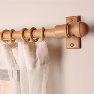 May include: A wooden curtain pole with a ball finial and wooden rings. The pole is mounted on a light-coloured wall. White sheer curtains hang from the rings, creating a soft, airy look. The pole is made of light-coloured wood.