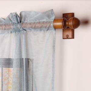 May include: A light blue, sheer curtain panel with a ruffled top, hanging from a wooden rod. The rod is supported by a wooden bracket with a ball finial. The curtain allows light to pass through. The rod and bracket are made of wood.