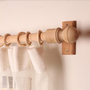 May include: Wooden curtain pole with a ball-shaped end and matching rings. The pole is mounted on a light-coloured wall. The curtains are white and semi-transparent, hanging from the rings.