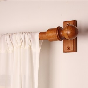 May include: A wooden curtain pole with a decorative finial and mounting bracket. The pole is a warm brown colour, and the finial is a rounded shape. A sheer white curtain hangs from the pole, creating a soft, airy look. The pole is mounted on a white wall.