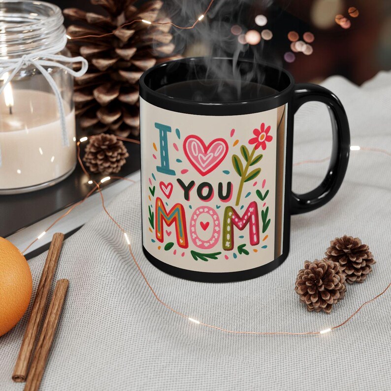 K&ouml;nnte beinhalten: Schwarze Kaffeetasse mit den Worten "I ❤️ YOU MOM" in farbenfrohen, skurrilen Buchstaben. Die Tasse hat einen schwarzen Henkel und Rand und ist mit Herzen und Blumen verziert. Eine Kerze, ein Tannenzapfen und Lichterketten befinden sich im Hintergrund.