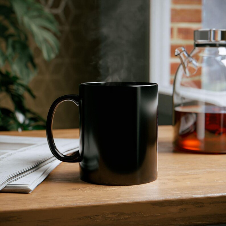 K&ouml;nnte beinhalten: Eine schwarze Keramiktasse mit gebogenem Henkel, die Dampf abgibt, steht auf einer Holzoberfl&auml;che. Im Hintergrund sind eine Glasteekanne mit einer rotbraunen Fl&uuml;ssigkeit und eine Zeitung zu sehen.