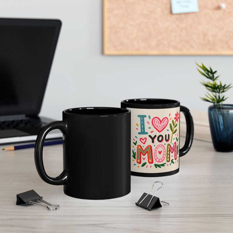 K&ouml;nnte beinhalten: Zwei schwarze Keramikbecher. Einer ist einfarbig schwarz, der andere hat einen cremefarbenen Innenraum und die Worte "I ❤️ YOU MOM" in farbenfrohen Buchstaben. Das Design umfasst ein rotes Herz, Blumen und dekorative Elemente.