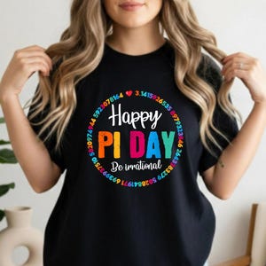 Op de afbeelding: Zwart T-shirt met een kleurrijke "Happy Pi Day" afbeelding. Het ontwerp bevat een cirkel van getallen en de zin "Be irrational". Het shirt wordt gedragen met een spijkerbroekshort.