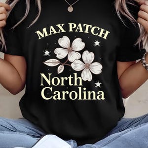 Peut inclure: T-shirt noir avec l'inscription "MAX PATCH North Carolina" en police serif, et un motif floral. Le motif comprend trois fleurs blanches avec des tiges et des feuilles marron, et des étoiles. Le t-shirt a un col rond.