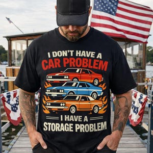 May include: Black t-shirt with the text "I DON'T HAVE A CAR PROBLEM I HAVE A STORAGE PROBLEM." The shirt features three classic cars in red, blue, and orange with flames. An American flag is in the background.