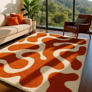 May include: A rectangular rug featuring an abstract design of orange shapes against a cream backdrop. The rug is in a living room with a sofa, armchair, and large windows offering views of a green landscape.