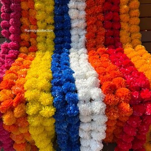 May include: A colourful display of flower garlands in shades of pink, orange, yellow, blue, white, and red. The garlands are arranged vertically, creating a visually striking and festive arrangement. The text "Ramsiyacollection" is visible.