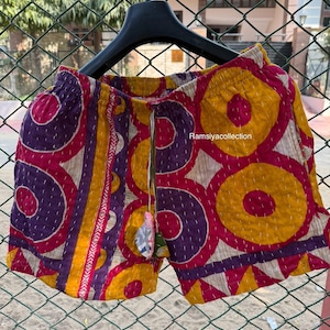 May include: Colorful shorts with a geometric pattern in yellow, purple, and pink. The shorts have a drawstring waist and are made of a patterned fabric. The shorts are hanging on a black hanger.