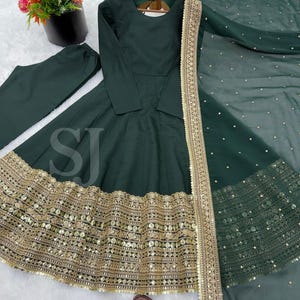 May include: A dark green Anarkali suit with gold embroidery and sequin embellishments. The set includes a long-sleeved dress, matching pants, and a sheer dupatta with gold trim. The dress features a flared skirt and intricate detailing along the hemline.