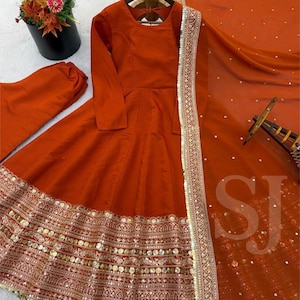 May include: An orange-red Anarkali suit with long sleeves, a flared skirt, and intricate gold embroidery along the hem. The ensemble includes matching pants and a sheer dupatta with sequin accents and a gold border. A wooden hanger supports the dress.