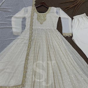 May include: A white, long-sleeved dress with intricate sequin detailing and gold trim. The dress features a round neckline with ornate gold embellishments and is paired with matching white pants and a sheer scarf.