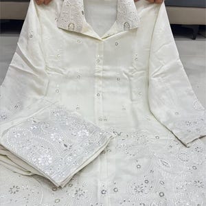 May include: Cream-coloured shirt and trousers set. The shirt has a collared neckline with embroidered detailing and small embellishments. The trousers have an embroidered border. The set is made of a soft, flowing fabric, suitable for warmer weather.
