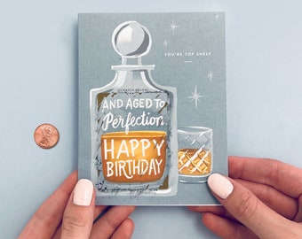 May include: A blue birthday card with a drawing of a decanter and a glass of whiskey. The text on the card reads "And aged to perfection. Happy Birthday." and "You're top shelf".