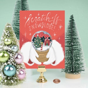 May include: A red card with the text "scratch-off snowglobe!" features a snow globe illustration held by white gloved hands. The snow globe shows a Christmas scene with a house, trees, and a candy cane. Miniature Christmas trees and ornaments surround the card.