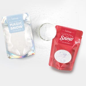 May include: Two pouches of Inklings brand artificial snow powder. The left pouch is iridescent with a light blue label reading Make Your Own Magic Snow. The right pouch is red with a label reading North Pole Snow. A small glass bowl of white powder sits between them.