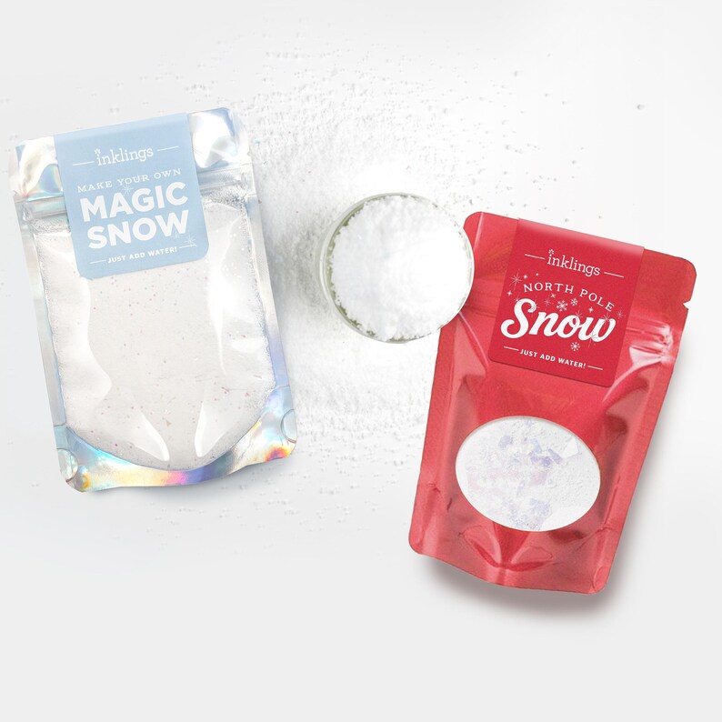 May include: Two pouches of Inklings brand artificial snow powder. The left pouch is iridescent with a light blue label reading Make Your Own Magic Snow. The right pouch is red with a label reading North Pole Snow. A small glass bowl of white powder sits between them.