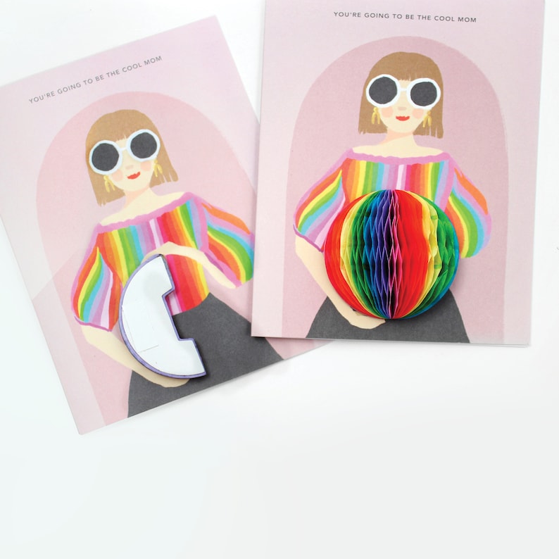 Op de afbeelding: Een kaart met een regenboogkleurig gestreept ontwerp en de tekst "YOU'RE GOING TO BE THE COOL MOM". De kaart toont een cartoonillustratie van een persoon die regenboogkleurige kleding en een zonnebril draagt en een regenboogkleurige pop-upbal vasthoudt.