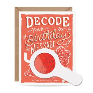 May include: A birthday greeting card with a red background and white text that reads "Decode Your Birthday Message." A white magnifying glass reveals a hidden message. The card is in a brown envelope.